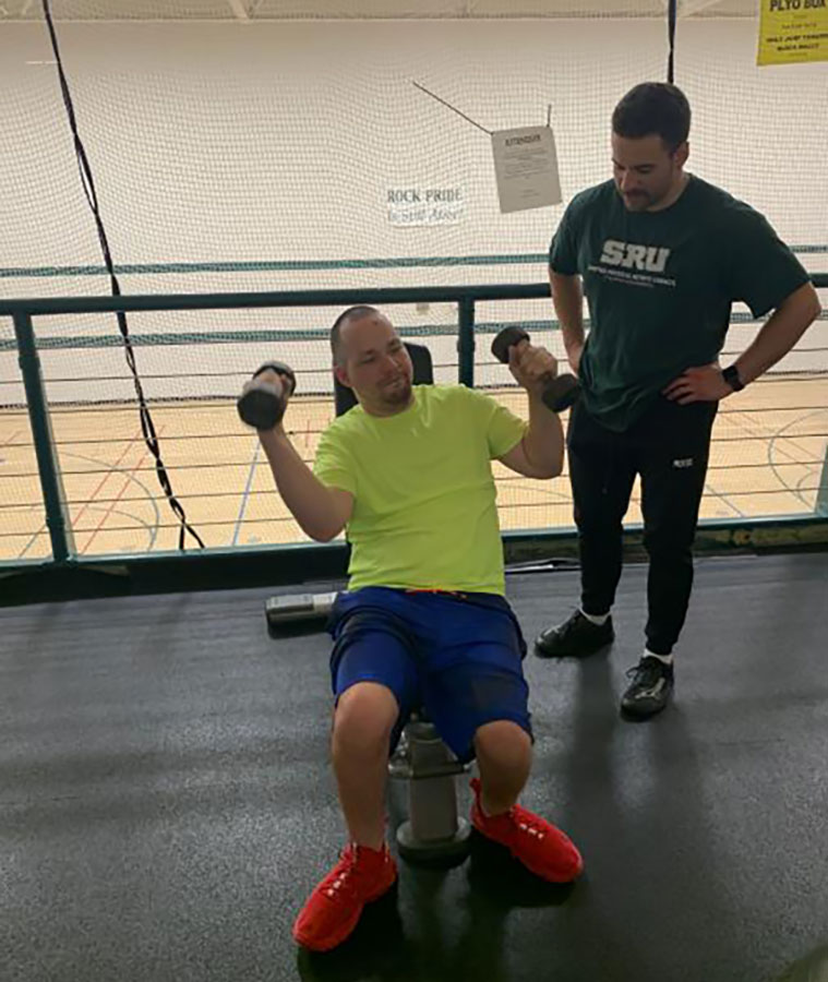 A coach supervising a seated member pressing dumbbells overhead